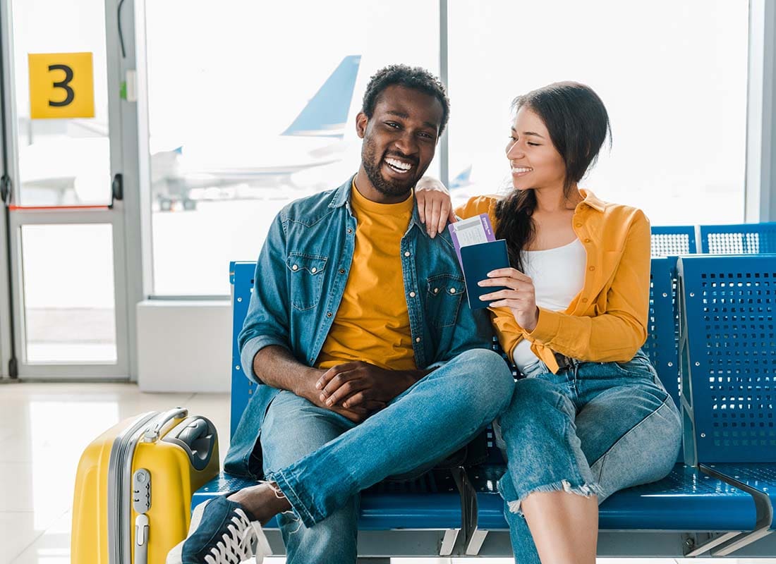 Travel Insurance - Smiling Young Couple Sitting in the Departure Lounge with Baggage and Tickets in the Airport with Airplane behind in the Distance