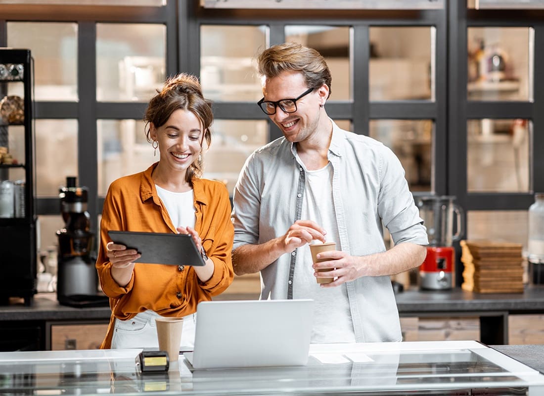 Business Insurance - Cheerful Young Female and Male Small Business Owners Standing Behind the Front Counter of Their Cafe Looking at a Tablet Together
