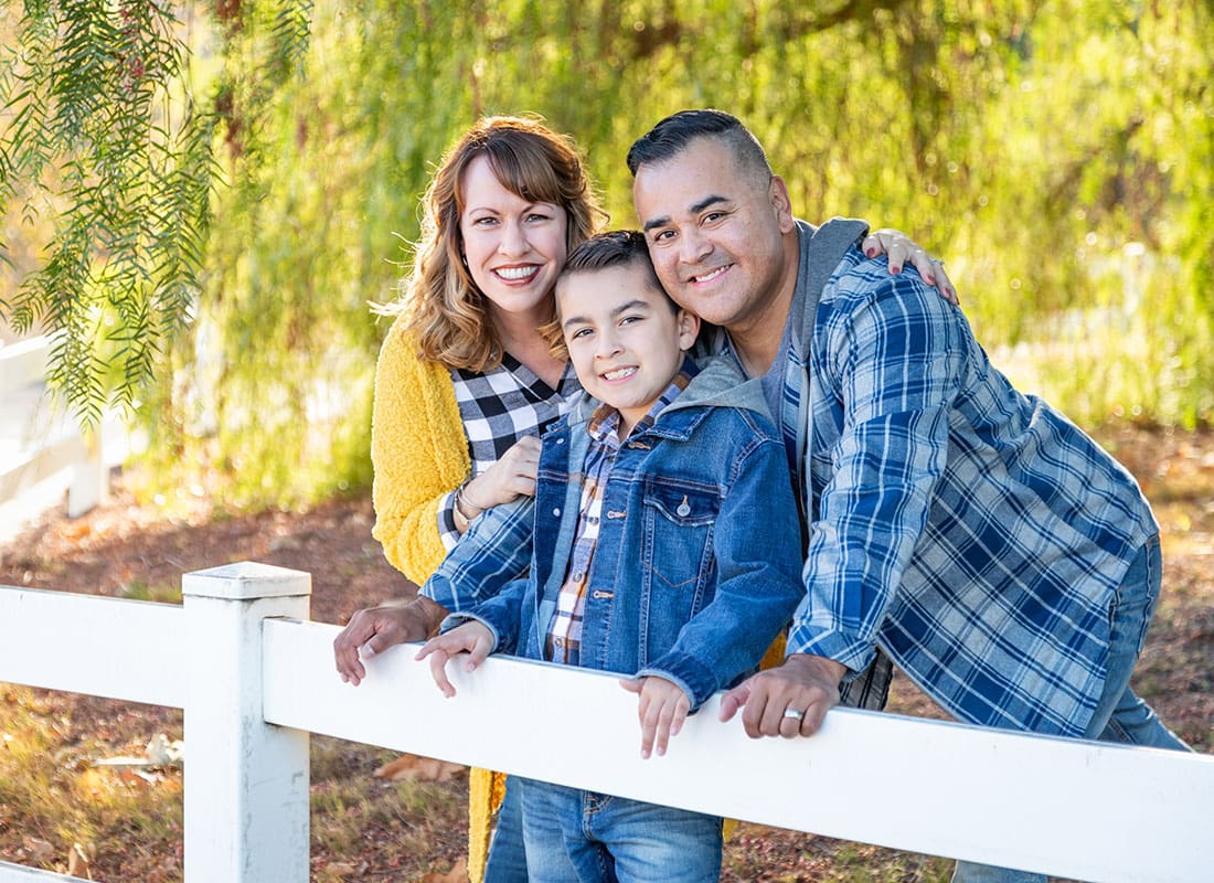 Personal Insurance - Portrait of a Cheerful Diverse Family with a Young Son Standing Under a Green Tree Next to a White Picket Fence on a Sunny Day